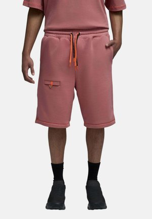 Homme portant un short rose poussiéreux jusqu'aux genoux avec des cordons orange, une poche latérale, des chaussettes noires et des chaussures de sport noires.