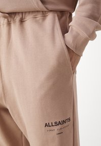 Béžové tepláky s rukou v kapse a černým logem "ALLSAINTS", které zahrnuje japonské znaky a nápis "LONDON" pod ním.