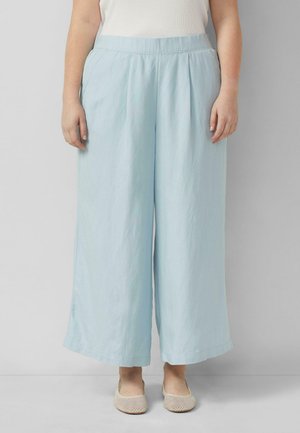 Persoon draagt lichtblauwe wijde broek met elastische tailleband en beige mesh ballerina's, staand tegen een effen grijze muur.