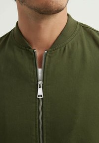 Veste bomber vert olive avec un col rond et une fermeture éclair en métal. Le tissu semble texturé avec une finition lisse.