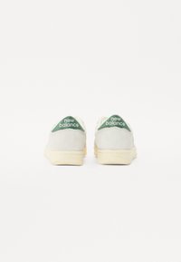 White suede sneakers with a rounded shape, featuring green branding accents on the back, and a beige rubber sole.