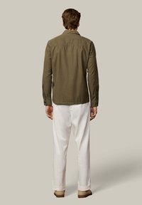 Chaqueta verde oliva con mangas largas, puños con botones y textura lisa, combinada con pantalones blancos y zapatos marrones, vista desde atrás.