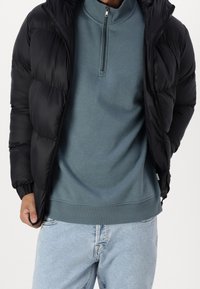 Musta puffatakki korkean kauluksen kanssa, jossa on tikattu muotoilu ja joustavat hihansuut, päällä teal-värinen vetoketjukauluksellinen collegepaita. Vaaleat farkut.