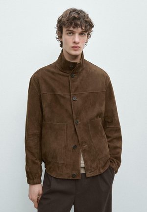 Junger Mann, der eine braune Wildlederjacke mit großen vorderen Taschen trägt, steht vor einem schlichten hellen Hintergrund.