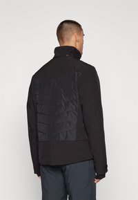 Schwarze Jacke mit hohem Kragen, ausgestattet mit einem quilted Paneel auf der Rückseite aus einem kontrastierenden matten Material. Glatte Textur, modernes Design.