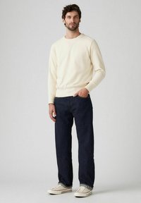 Sudadera color crema con un diseño de cuello en V, combinada con jeans de mezclilla azul oscuro y zapatillas de color claro, creando un atuendo casual.