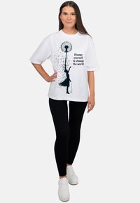 Wit katoenen t-shirt met oversized ontwerp. De afbeelding toont een meisje dat een paardenbloem vasthoudt, met de tekst: "Verander jezelf om de wereld te veranderen."