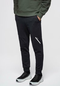 Mand iført sorte, tilspidsede joggerbukser med hvid diagonal stribe og sorte og hvide sneakers, hænderne i lommerne.