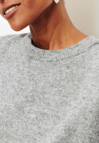 Pull gris à maille texturée, orné de sequins argentés dispersés. Gros plan sur l'encolure côtelée et le tissu doux et scintillant.