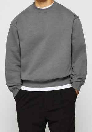 Mężczyzna nosi szarą bluzę crewneck na białej koszuli, czarne spodnie, ręce swobodnie w kieszeniach, na jednolitym tle.