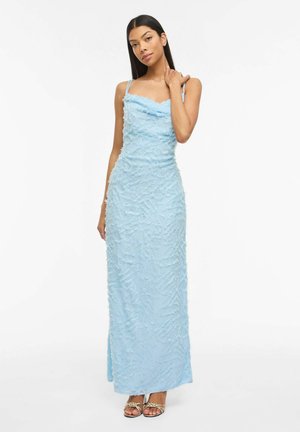 Ženska z dolgimi temnimi lasmi, oblečena v brezrokavno svetlo modro teksturirano maksi obleko in z zlatimi sandali z naramnicami, stoji z eno roko na vratu.