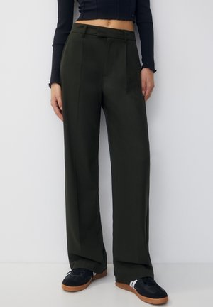 Modell trägt dunkelgrüne, weit geschnittene Hosen, ein schwarzes, langärmeliges, kurzes Oberteil und schwarze Sneaker mit braunen Sohlen und weißem Streifendetail.