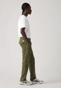 Olive green pants, relaxed fit, with side pockets. Paired with a plain white t-shirt and silver athletic shoes. Smooth fabric texture.