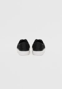 Paire de baskets basses noires en mesh avec semelles blanches vues de dos sur un fond clair uni.