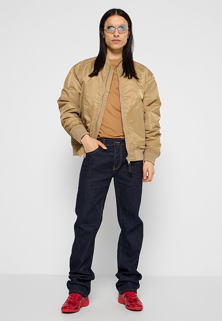 Beige Bomberjacke mit gerippten Bündchen, einem braunen T-Shirt darunter, dunklen Jeans und roten Turnschuhen, getragen mit einer Brille mit klarem Rahmen.