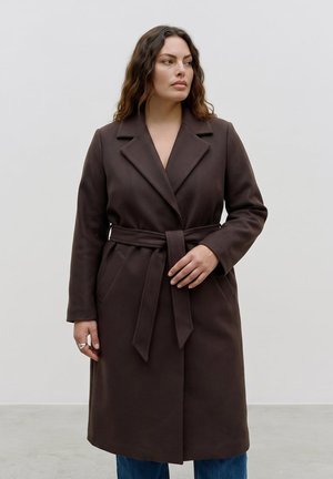 Femme aux cheveux noirs ondulés portant un manteau marron ceinturé et un jean bleu, debout sur un fond blanc uni, regardant sur le côté.