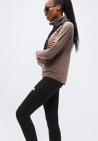 Καλοσιδερωμένο καφέ μπουφάν με φερμουάρ και ριμπ ύφανση· συνδυασμένο με μαύρα κολάν που φέρουν λευκό λογότυπο. Το μοντέλο φοράει γυαλιά ηλίου και στέκεται με τα χέρια σταυρωμένα.