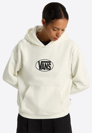 Person iført en off-white Vans hættetrøje med sort logo, kiggende ned, hænder bag ryggen og klædt i sorte bukser.