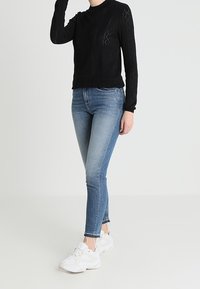 Černý pletený svetr s propracovaným vzorem výřezů, doplněný světle modrými úzkými džíny a bílými teniskami. Oblečení má přiléhavý střih.