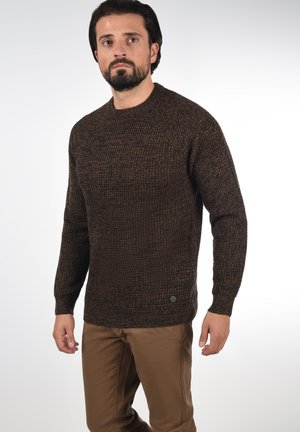 Bruine gebreide trui met een textuurpatroon, ronde hals, lange mouwen en een klein merketiketje aan de onderkant. Gecombineerd met beige broek.
