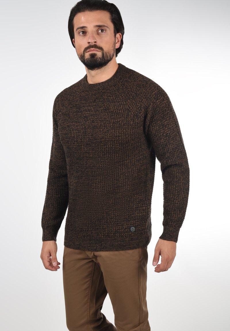 Brauner Strickpullover mit strukturiertem Muster, Rundhalsausschnitt, langen Ärmeln und einem kleinen Logo-Tag am unteren Rand. Kombiniert mit beigen Hosen.
