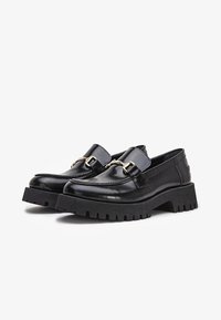 Mocassins en cuir noir avec semelles à crampons épaisses et détail en métal doré en forme de mors sur le dessus, présentés sur un fond blanc uni.