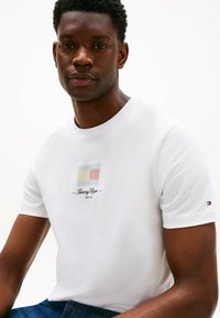 Hvid bomulds t-shirt med et farverigt logo i blå, gul og rød striber. Rund halsudskæring med korte ærmer og et lille logo på ærmet.