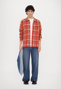 Rdeča in rumena karirana srajca, zakopčana, nosi se čez belo srajco. Modri široki kavbojki in črne čevlje. Drži svetlo modro jeans jakno.