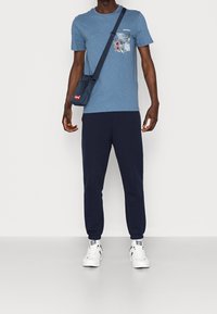 Modré tričko s květinovým vzorem na kapse, námořnické tepláky, bílé tenisky a malá modrá taška přes rameno s červeným logem.