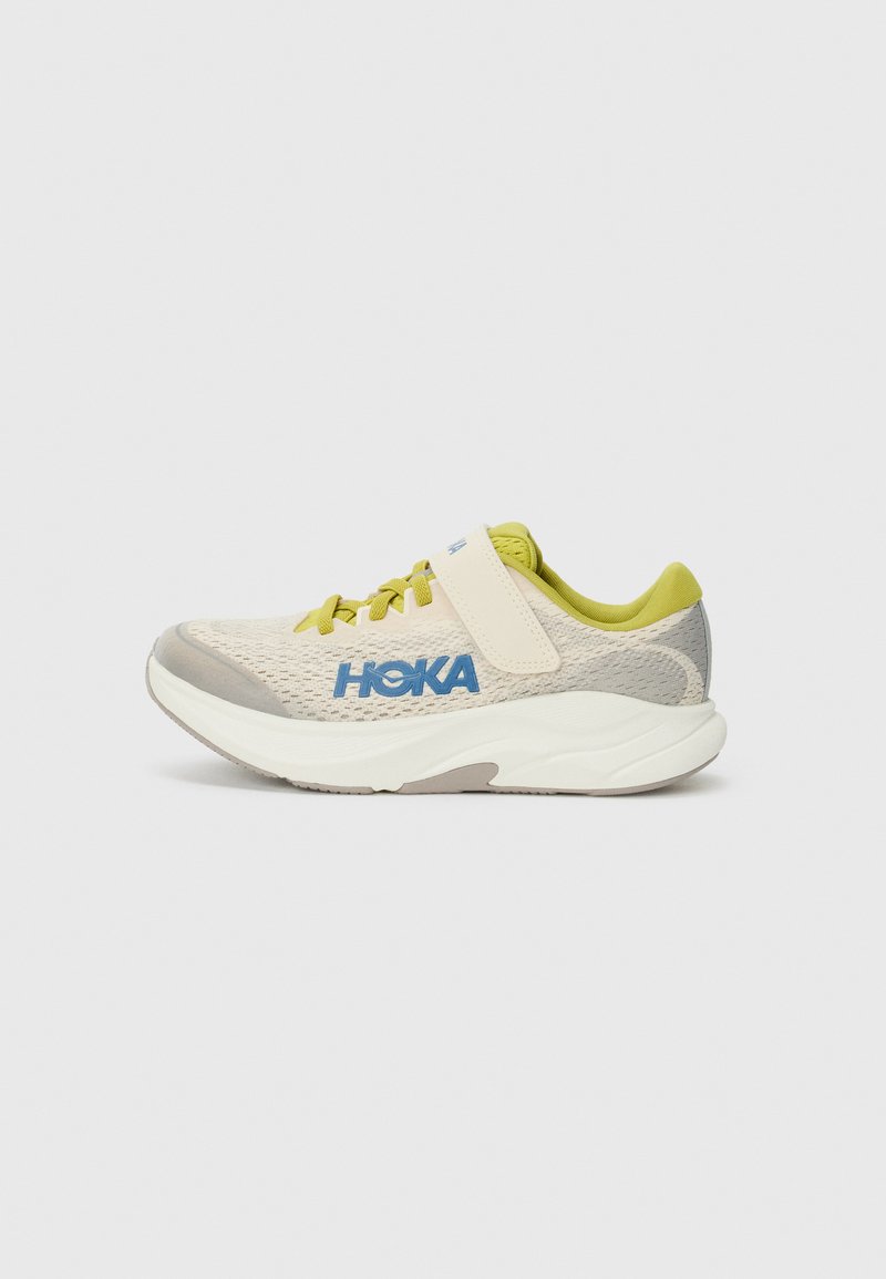Beige athletic shoe with mesh upper, green accents, and blue logo. Features laces and adjustable strap, thick white midsole, and textured gray outsole.