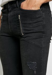 Jeans negros de corte Slim, con bolsillos con cremallera, detalles desgastados y una textura suave. La tela es denim con un ligero desvanecimiento.