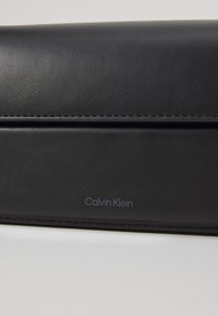 Černá kožená kabelka s hladkou texturou, s kulatým tvarem a jemným prošíváním. Logo 'Calvin Klein' vytištěné šedě.