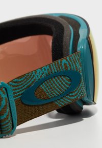 Skidglasögon med en tealram, runda linser och ett texturerat brunt band med ett vågmönster. Slät yta och bekväm skumfoder.
