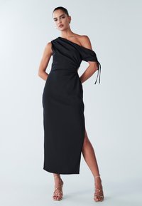 Robe noire à une épaule avec un corsage ajusté, fente sur le côté et texture douce ; détail noué sur la manche. Accentuée par des talons dorés à lanières.