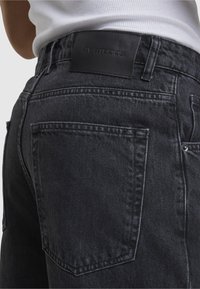 Prim-plan al buzunarului din spate al blugilor de culoare gri închis și al patch-ului de brand din piele neagră de pe talie, purtați cu un top alb cu dungi.