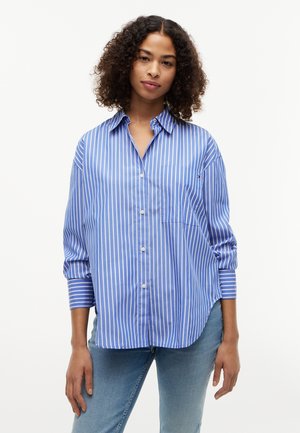 Femme aux cheveux bouclés portant une chemise à boutons rayée verticalement bleu et blanc et un jean bleu clair, debout devant un fond blanc uni.