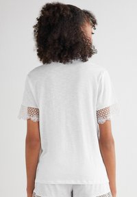 Witte t-shirt met korte mouwen, featuring kantaccenten op de mouwen, een ronde hals en zachte, gestructureerde stof. De achterkant toont de kantdetails duidelijk.