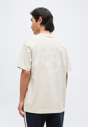 Homme aux cheveux noirs portant un t-shirt beige oversize avec un graphisme blanc pâle et un texte au dos, associé à un pantalon foncé avec des bandes blanches.