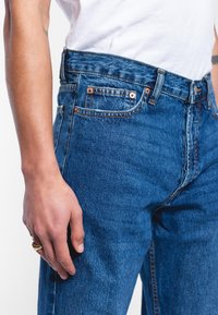 Vaqueros de mezclilla azul con cintura alta, diseño de cinco bolsillos, remaches de cobre y un acabado ligeramente deslavado, combinados con una camiseta blanca.