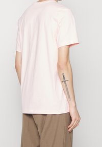 Camiseta de algodón de color rosa claro con cuello redondo y mangas cortas, que muestra una textura suave y un ajuste relajado. Se ven pantalones marrones abajo.