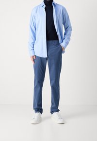 Camicia azzurro chiaro con bottoni sopra un dolcevita nero, abbinata a pantaloni blu e sneakers bianche. Tessuto liscio e vestibilità su misura.