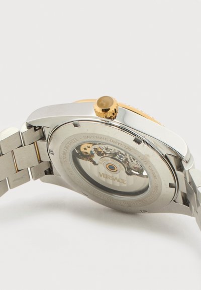 Sidabrinės ir auksinės spalvos nerūdijančio plieno laikrodžio nugarėlė, rodanti atvirą mechaninį judesį ir „Versace“ ženklinimą per safyro kristalą.