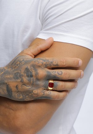 Gold ring with a rectangular red stone, worn on a tattooed hand, resting on a bare arm. White t-shirt background.