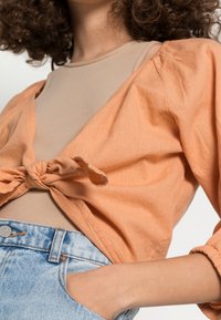 Top avvolgente arancione chiaro con maniche arricciate, legato davanti. Indossato sopra una camicia beige aderente, abbinato a jeans in denim blu a vita alta.