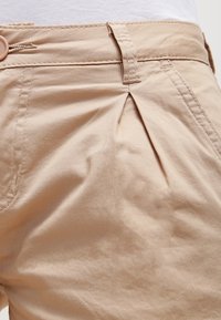 Vaaleat puuvillahousut, joissa on poimutettu etuosa, napitus ja vyölenkit. Kankaan pinta on sileä ja saumat siistit.