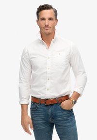 Camisa blanca de botones con cuello y bolsillo en el pecho, combinada con jeans azules y un cinturón marrón. Reloj en la muñeca, ajuste casual, mangas largas.