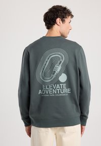 Felpa verde scuro con una stampa grafica sul retro caratterizzata da linee circolari, testo che recita "Elevate Adventure" e un accento rotondo.