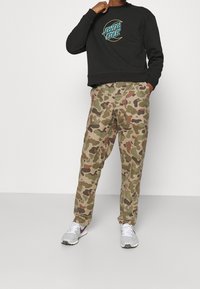 Black sweatshirt with "Santa Cruz" logo, paired with tan camouflage pants and white sneakers with purple accents. Casual fit, cotton fabric.