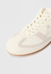 Superga v kremni barvi s gladkim usnjenim zgornjim delom in semiša, z vezalkami in ukrivljeno gumijasto podplatom. Na strani je natisnjen logotip.