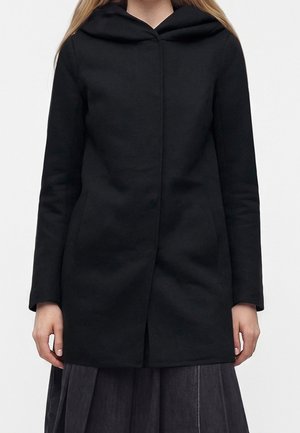 Femme portant un manteau noir à capuche avec manches longues et fermeture avant cachée, associé à une jupe plissée foncée.
